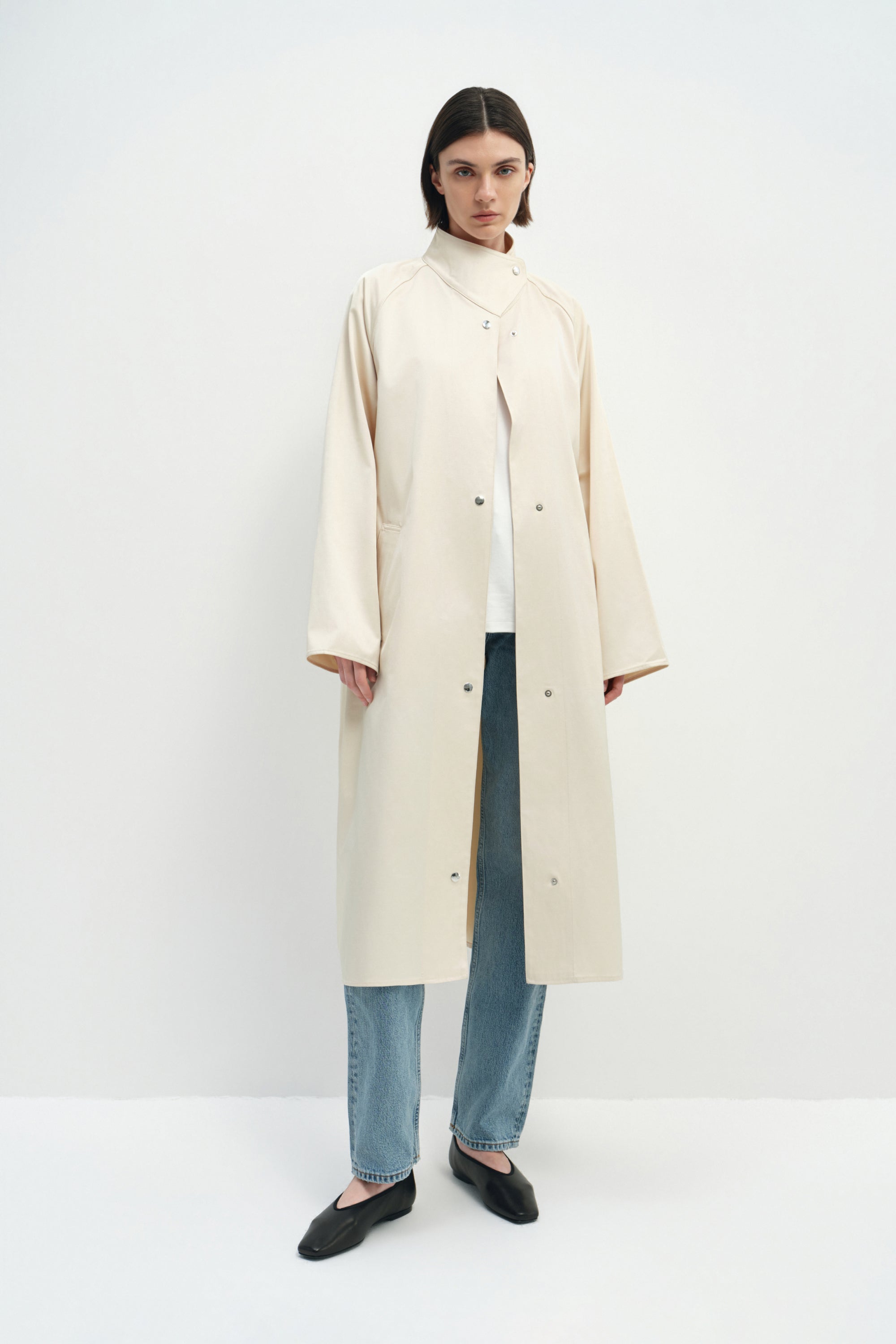 A model wearing the Rue Sophie Anne Satin Coat in crème, featuring a high stand collar, metallic snap-front closure, raglan sleeves, side pockets, and a relaxed longline silhouette, front view.