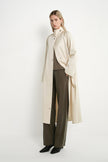 A woman models a long cream-colored coat over a brown top and dark olive wide-leg trousers, standing against a plain white studio background.