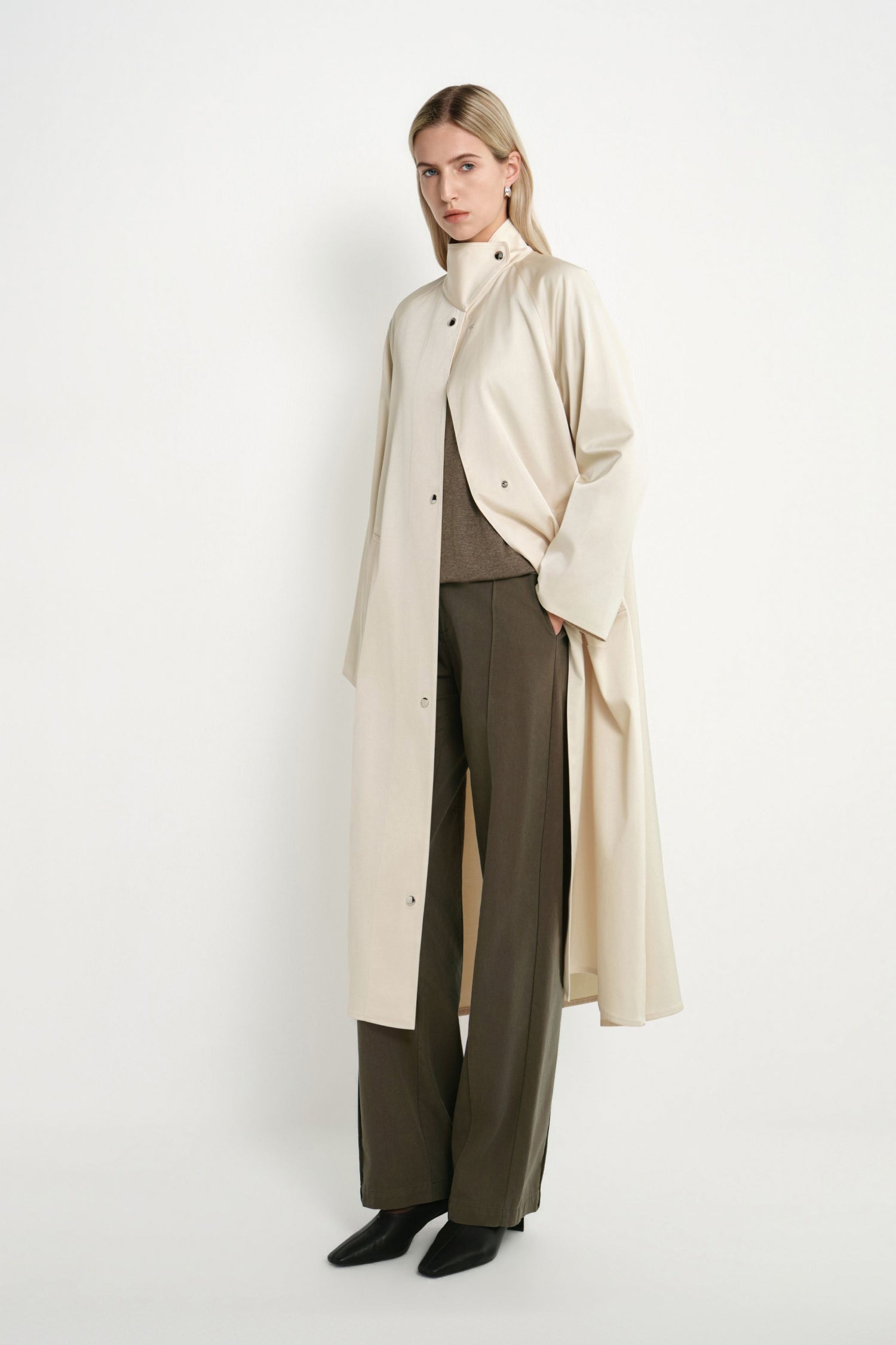 A woman models a long cream-colored coat over a brown top and dark olive wide-leg trousers, standing against a plain white studio background.