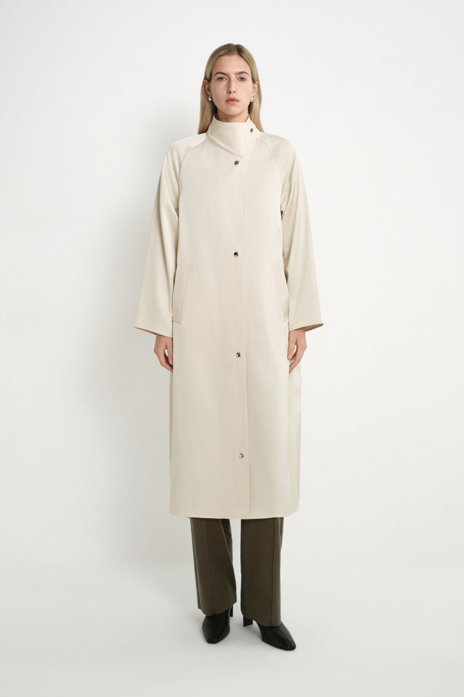 A model stands against a plain light gray studio background wearing a long cream-colored coat with a high mock neck, front buttons, and wide sleeves, paired with dark olive pants and black shoes.