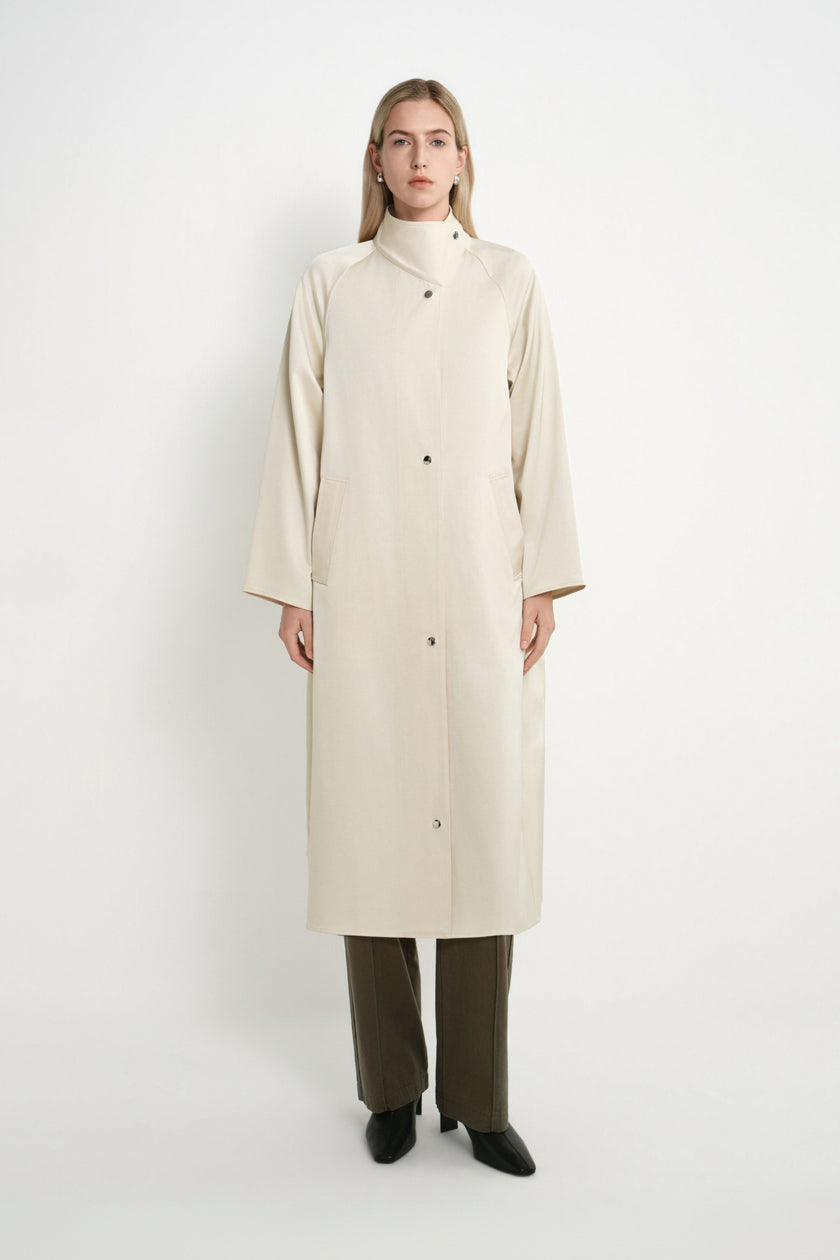 A model stands against a plain light gray studio background wearing a long cream-colored coat with a high mock neck, front buttons, and wide sleeves, paired with dark olive pants and black shoes.