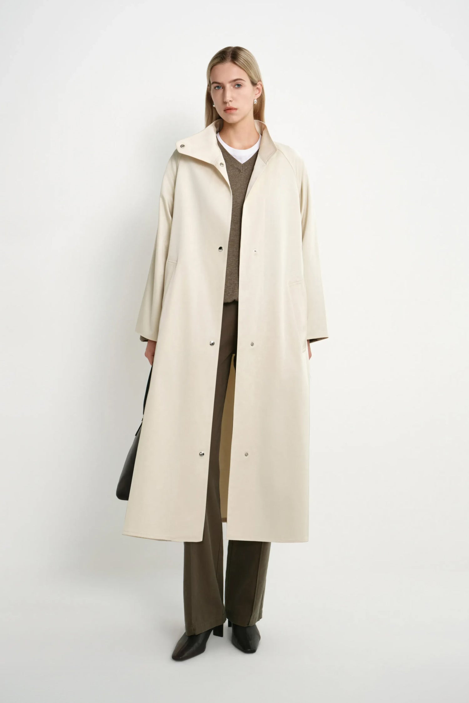 A woman stands against a plain light gray studio background wearing a long cream-colored coat over a brown sweater and brown wide-leg pants, with black shoes and a black handbag.