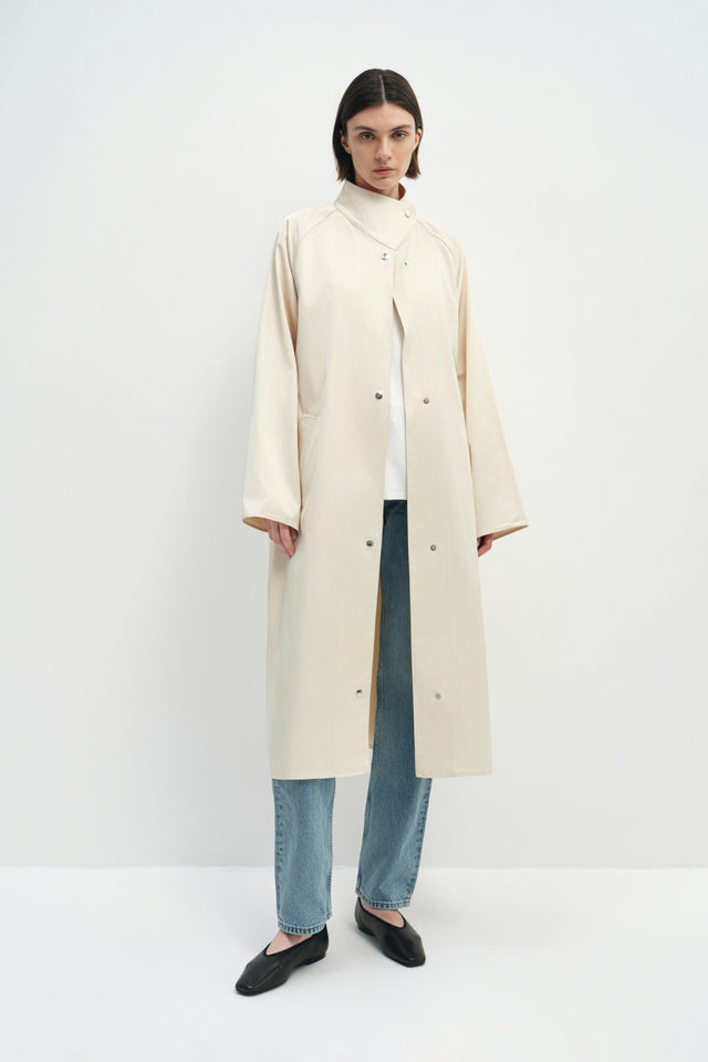 A model wearing the Rue Sophie Anne Satin Coat in crème, featuring a high stand collar, metallic snap-front closure, raglan sleeves, side pockets, and a relaxed longline silhouette, front view.