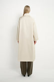 A model viewed from the back wearing a long, straight, off-white coat and wide-leg olive green trousers, standing against a plain white studio background.