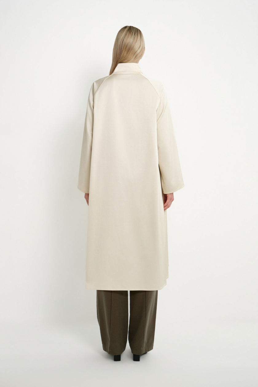 A model viewed from the back wearing a long, straight, off-white coat and wide-leg olive green trousers, standing against a plain white studio background.