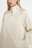 A close-up of a model wearing a cream-colored, high-neck long-sleeve jacket with silver snap buttons, set against a plain light background.