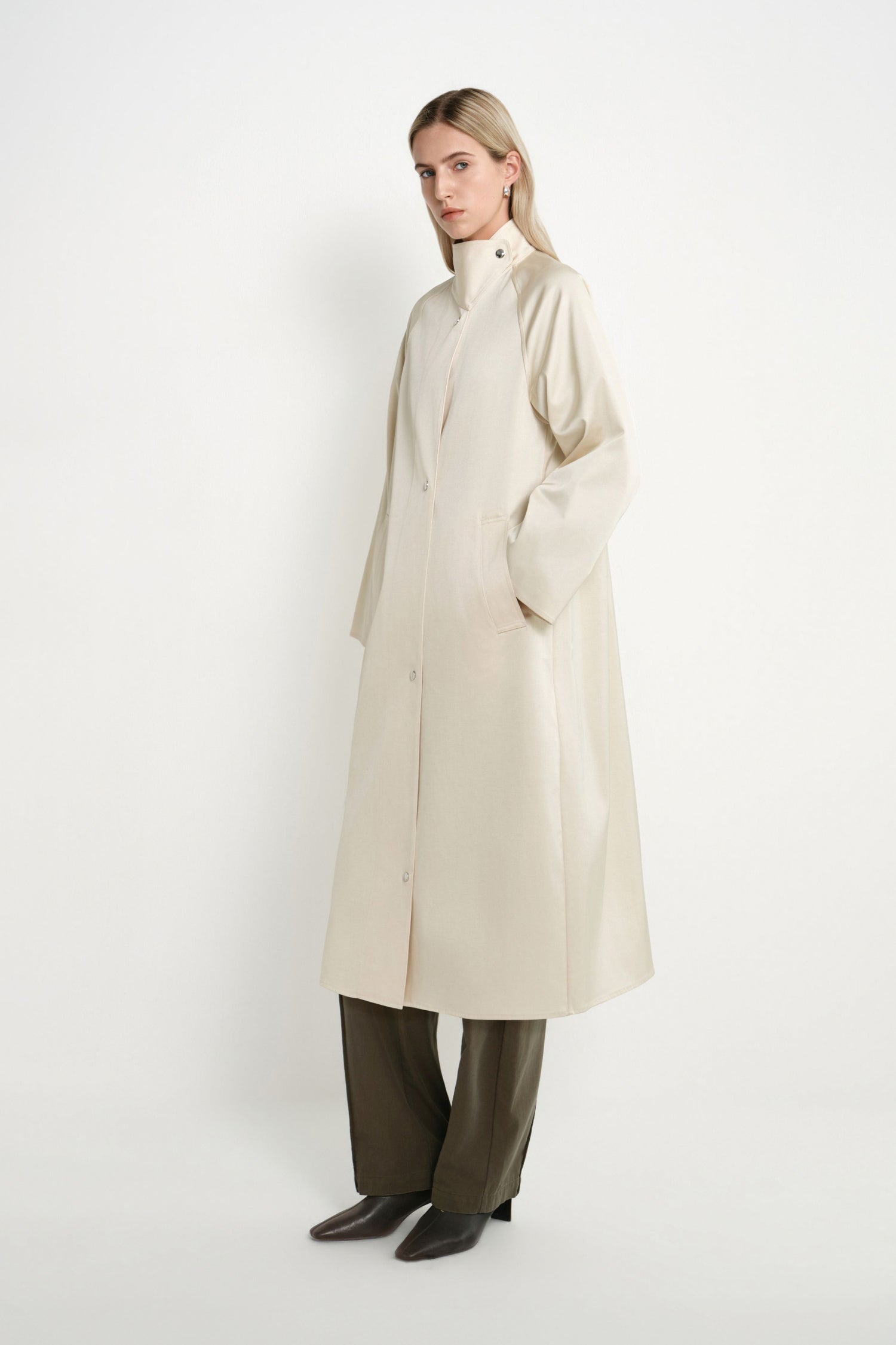 A woman modeling a long, high-neck, oversized cream-colored coat with wide sleeves, paired with dark olive wide-leg trousers and black pointed shoes, against a plain white studio background.