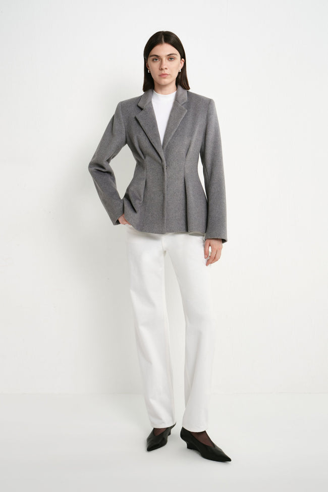 A model wearing the Rue Sophie Trenton Pleat Waist Wool Blazer in charcoal with white trousers, featuring notched lapels, structured shoulders, and a sculpted pleat waist, full front view.