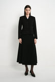 A model stands against a plain light gray studio background wearing a black tailored jacket with front buttons and a matching black midi skirt, paired with black tights and pointed black shoes.