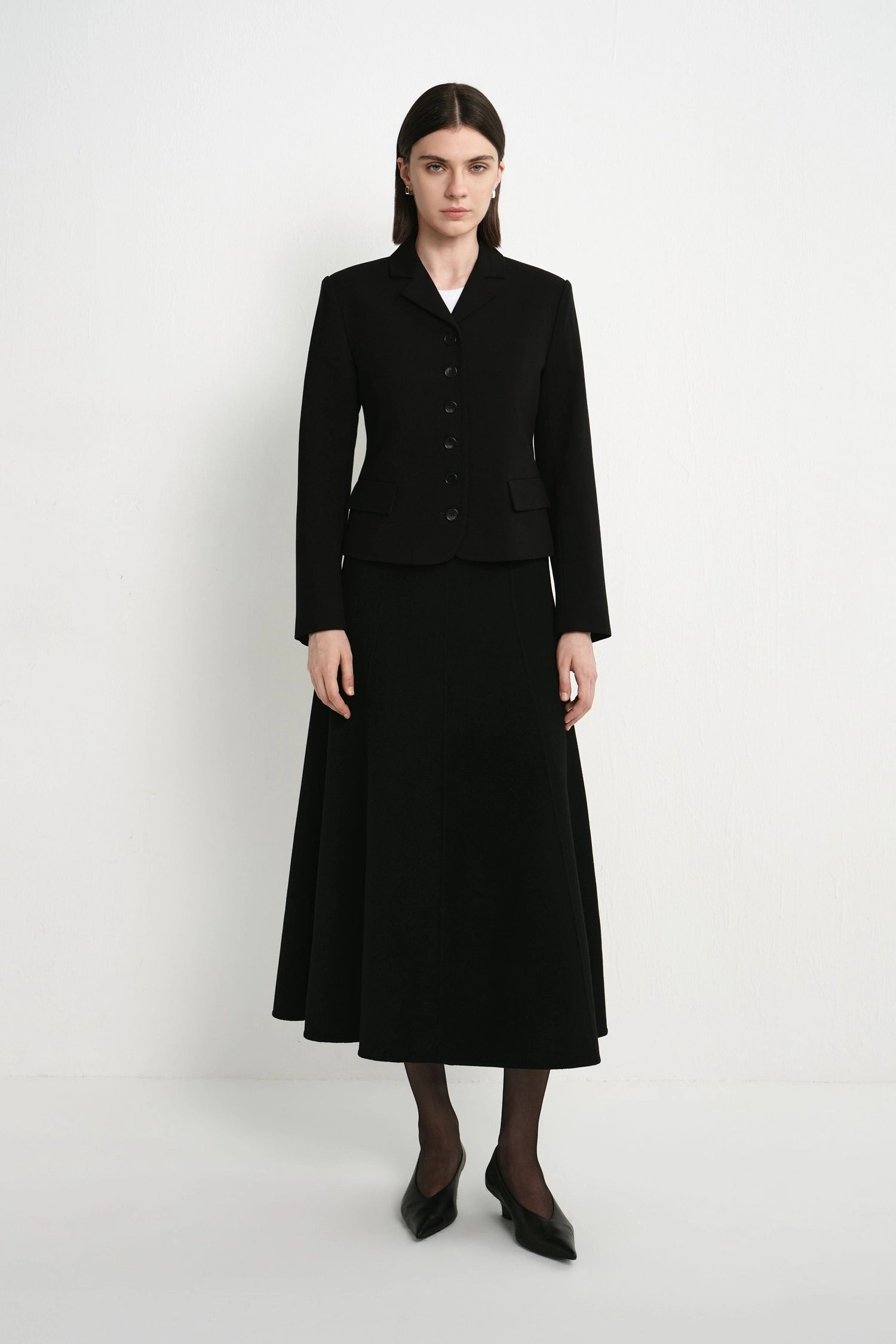A model stands against a plain light gray studio background wearing a black tailored jacket with front buttons and a matching black midi skirt, paired with black tights and pointed black shoes.
