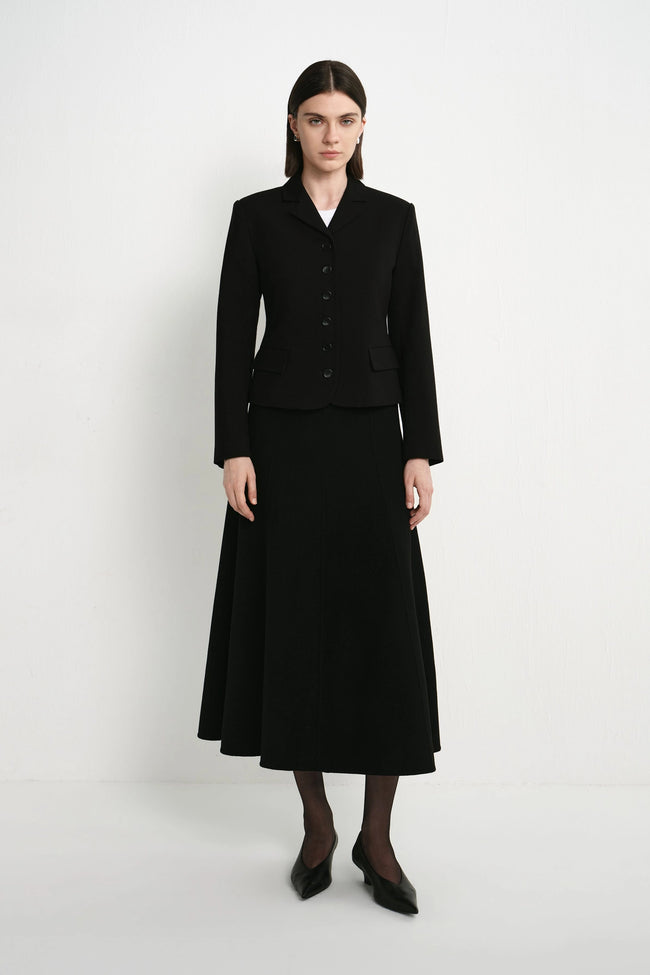 A model stands against a plain light gray studio background wearing a black tailored jacket with front buttons and a matching black midi skirt, paired with black tights and pointed black shoes.