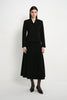 A model stands against a plain light gray studio background wearing a black tailored jacket with front buttons and a matching black midi skirt, paired with black tights and pointed black shoes.