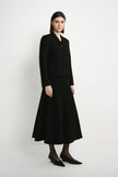 A model stands in a studio wearing a black tailored jacket with long sleeves and a matching black midi skirt. The outfit is styled with black tights and black pointed flats against a plain light gray background.