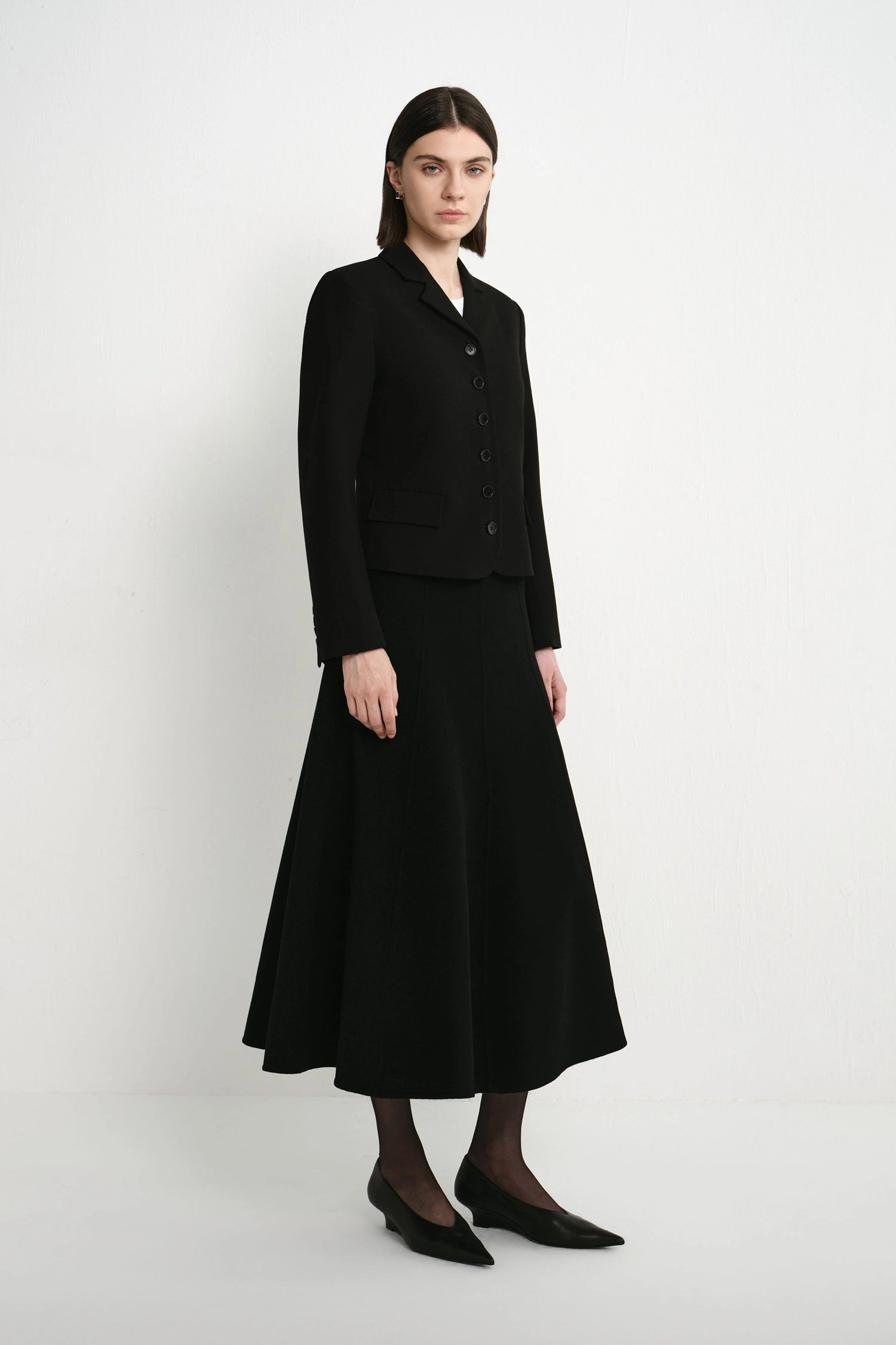 A model stands in a studio wearing a black tailored jacket with long sleeves and a matching black midi skirt. The outfit is styled with black tights and black pointed flats against a plain light gray background.