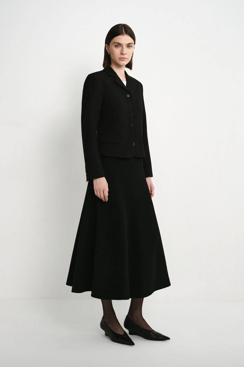 A model stands in a studio wearing a black tailored jacket with long sleeves and a matching black midi skirt. The outfit is styled with black tights and black pointed flats against a plain light gray background.