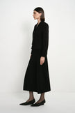 A woman stands in profile wearing a black tailored jacket with matching long skirt and black pointed flats against a clean white studio background.