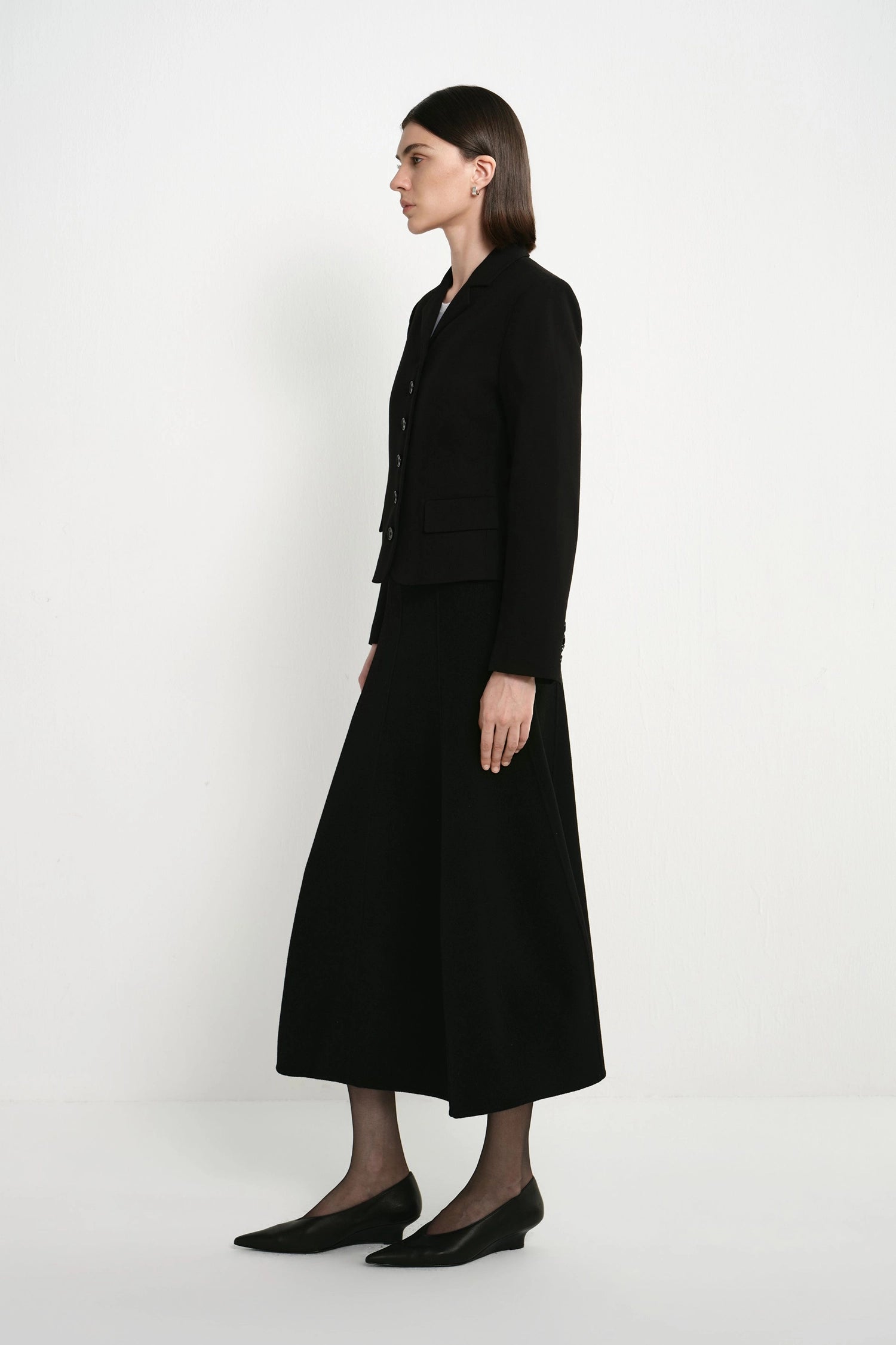 A woman stands in profile wearing a black tailored jacket with matching long skirt and black pointed flats against a clean white studio background.