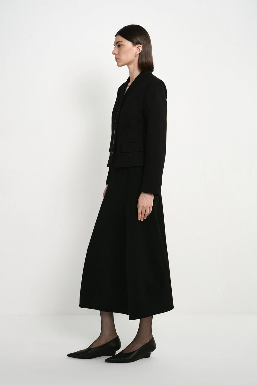 A woman stands in profile wearing a black tailored jacket with matching long skirt and black pointed flats against a clean white studio background.
