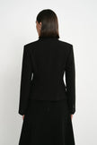 A woman viewed from the back wearing a structured black blazer and a long black skirt against a plain light gray studio background.
