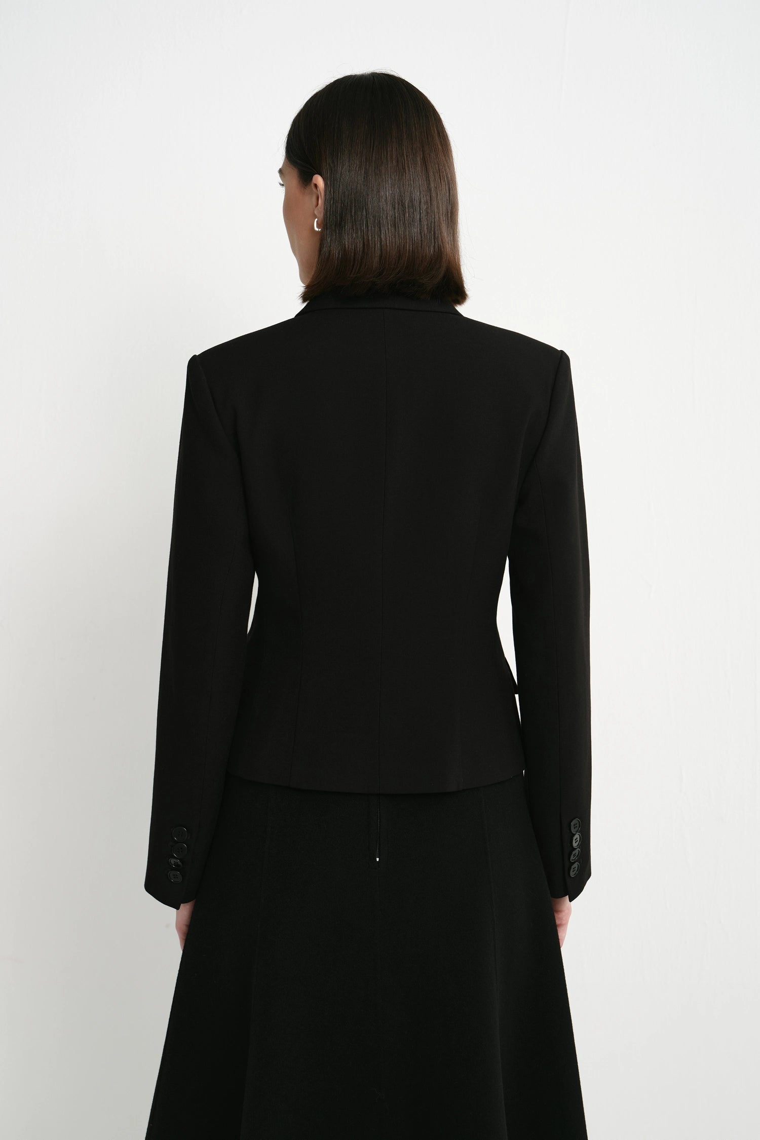 A woman viewed from the back wearing a structured black blazer and a long black skirt against a plain light gray studio background.