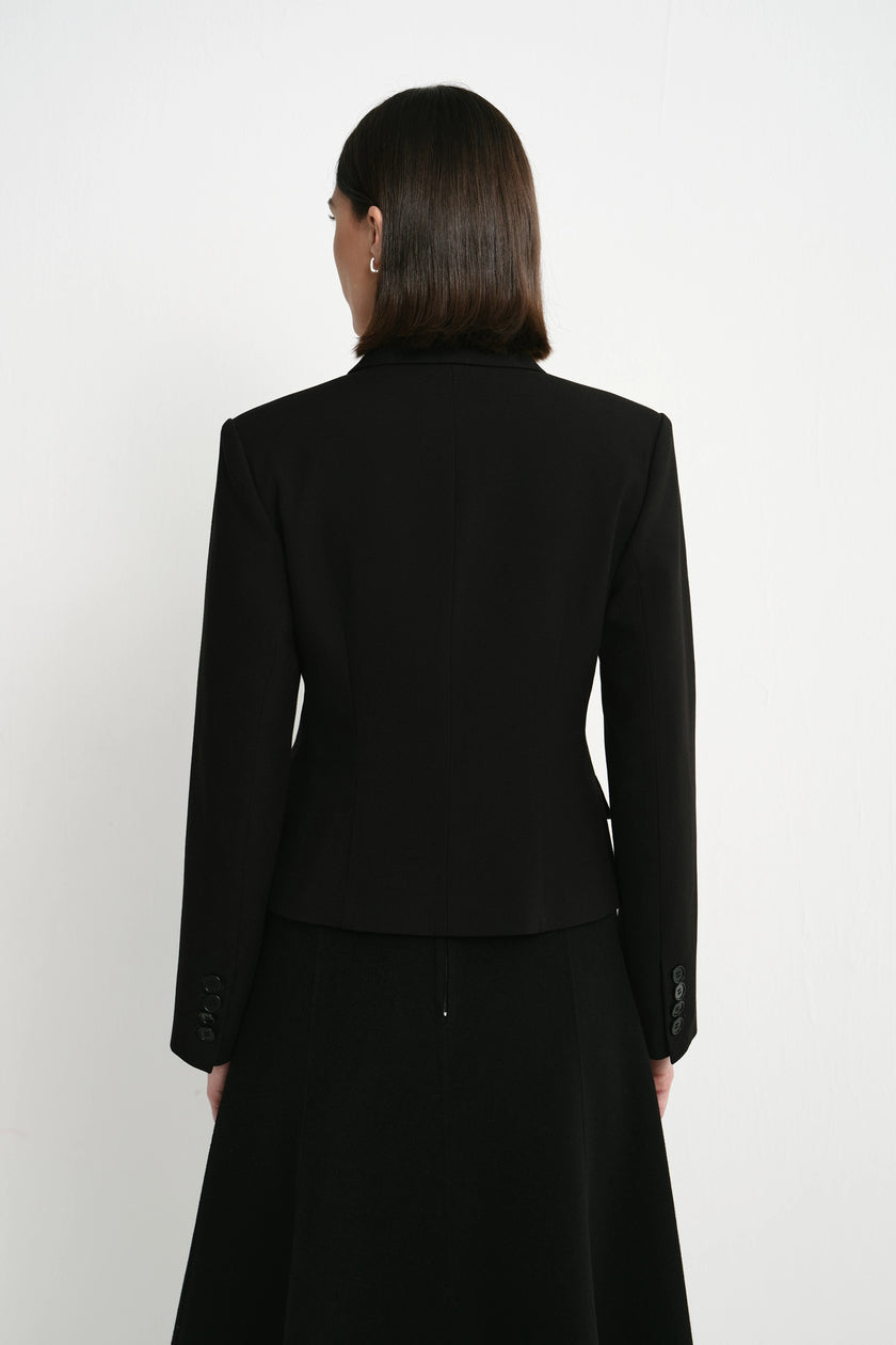 A woman viewed from the back wearing a structured black blazer and a long black skirt against a plain light gray studio background.