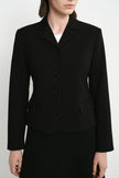 A model wearing a black tailored blazer with a notched lapel, five front buttons, flap pockets, and long sleeves, paired with a black skirt against a light gray studio background.