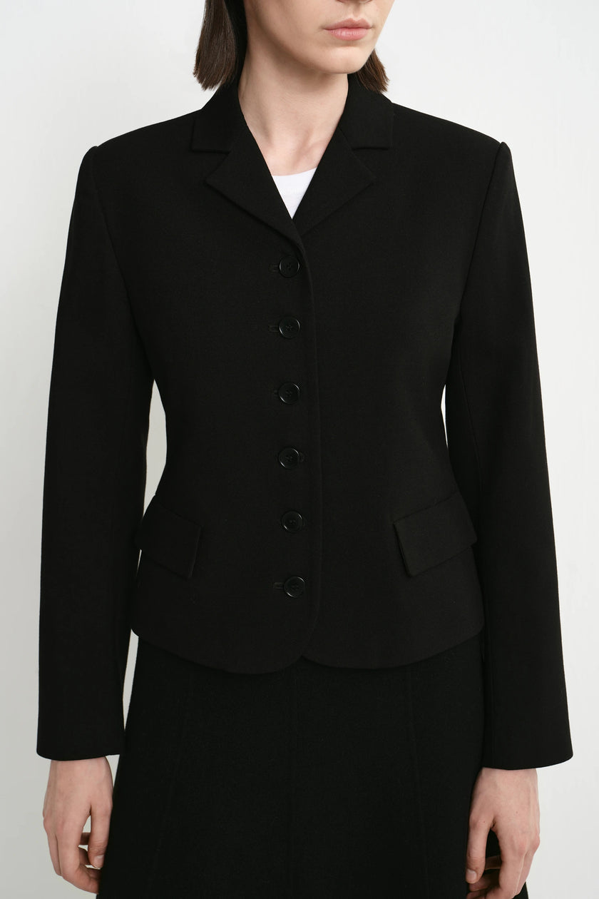 A model wearing a black tailored blazer with a notched lapel, five front buttons, flap pockets, and long sleeves, paired with a black skirt against a light gray studio background.
