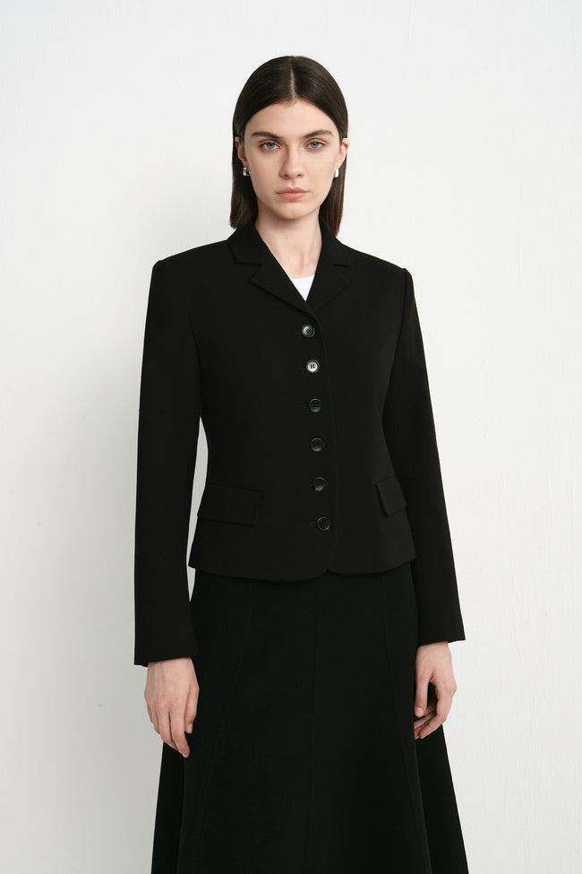 A model wearing a black tailored blazer with a row of dark buttons and a matching black skirt, standing against a plain light gray studio background.