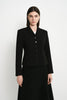A model wearing a black tailored blazer with a row of dark buttons and a matching black skirt, standing against a plain light gray studio background.