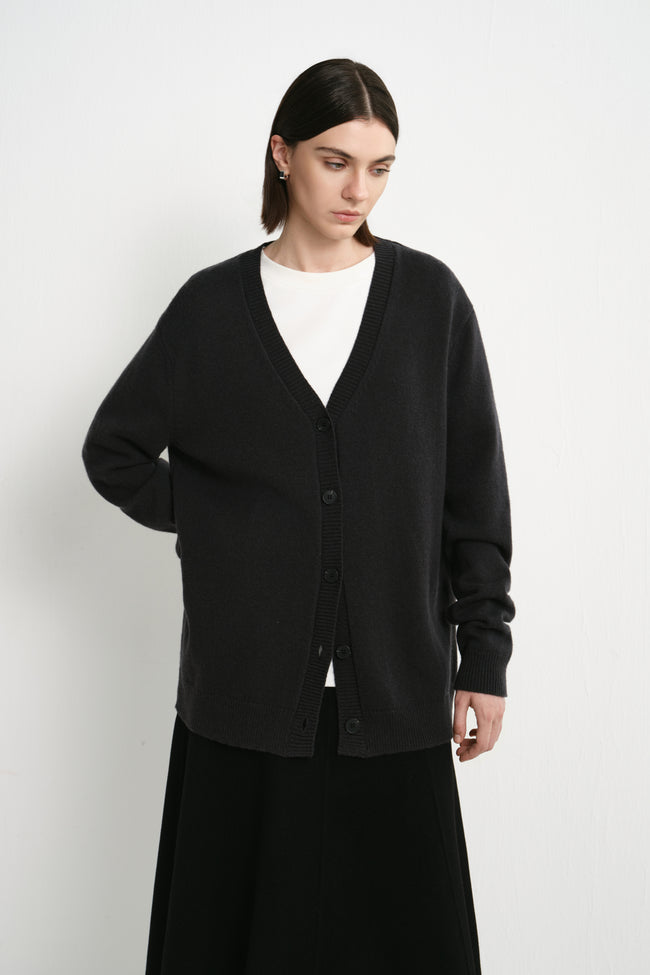 A model wearing the Rue Sophie Siho Cardigan Sweater in charcoal with a deep V-neckline, tonal buttons, long sleeves, and a relaxed oversized fit, three-quarter front view.