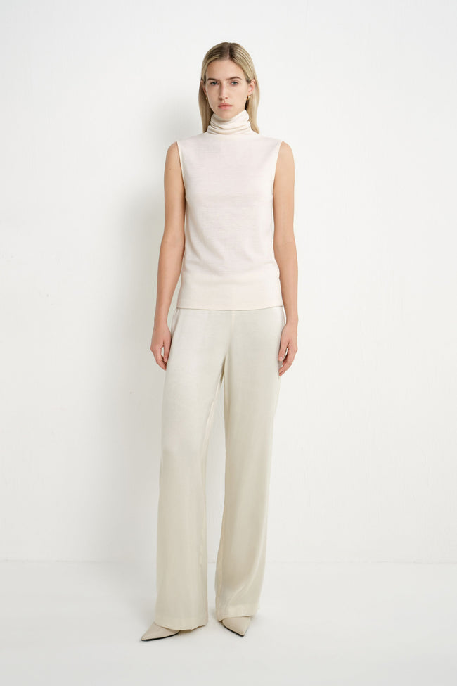 A model stands against a plain white studio background wearing a sleeveless cream turtleneck sweater and matching wide-leg trousers.