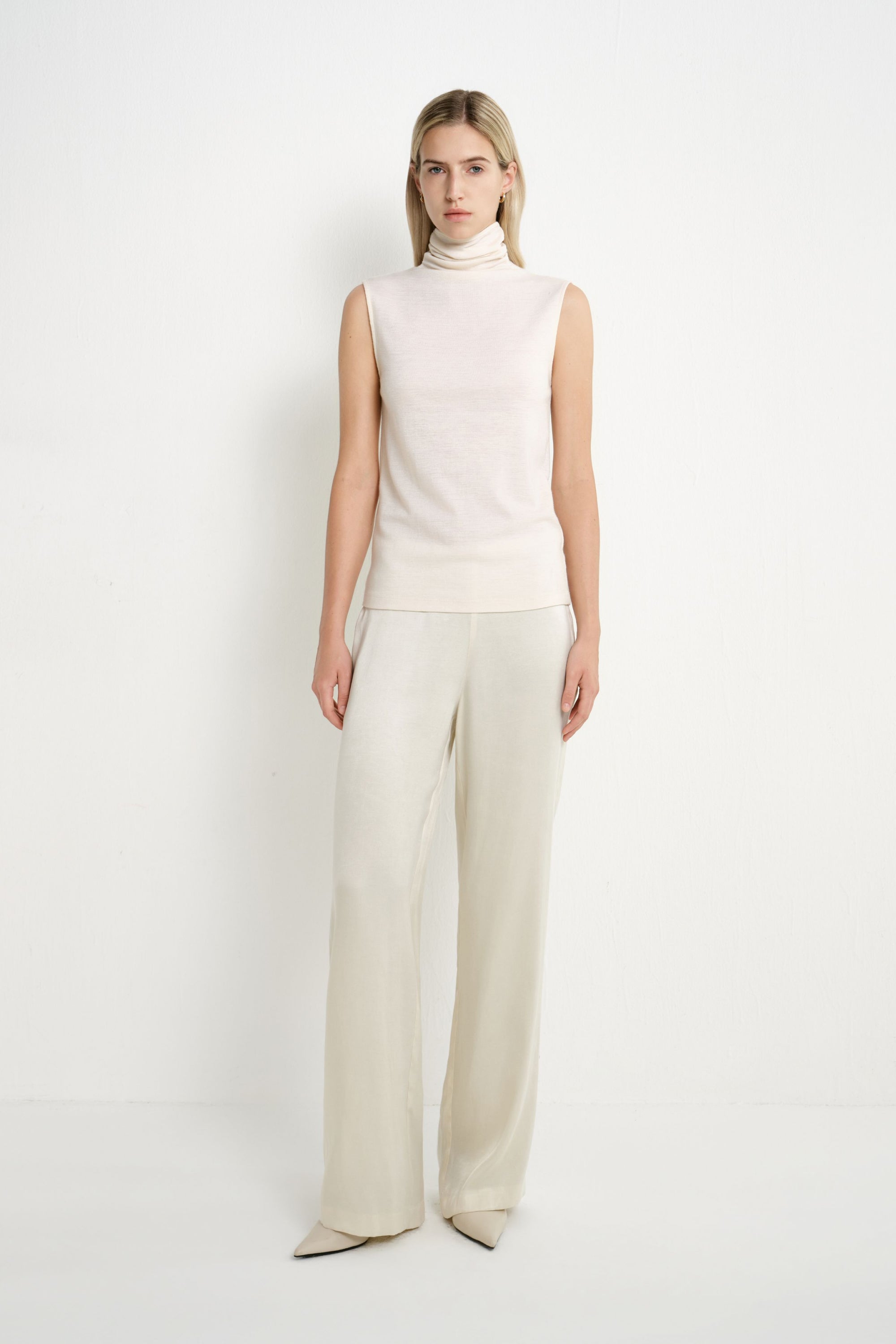 A model wearing a Rue Sophie creme sleeveless mock-neck top with a softly draped high neckline and clean straight silhouette, front view.