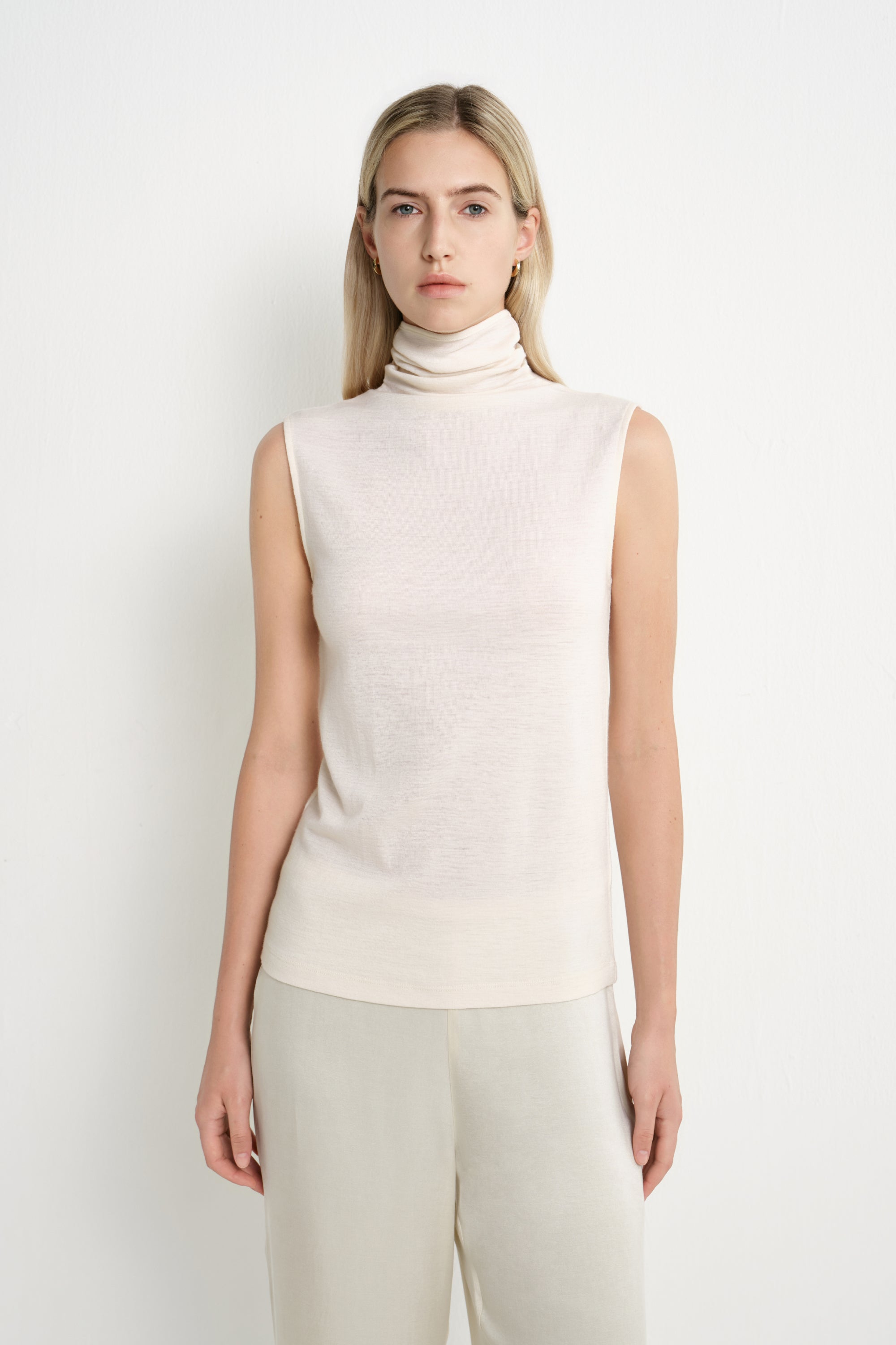 A model wearing a Rue Sophie creme sleeveless mock-neck top with a softly draped high neckline and clean shoulder construction, front view.
