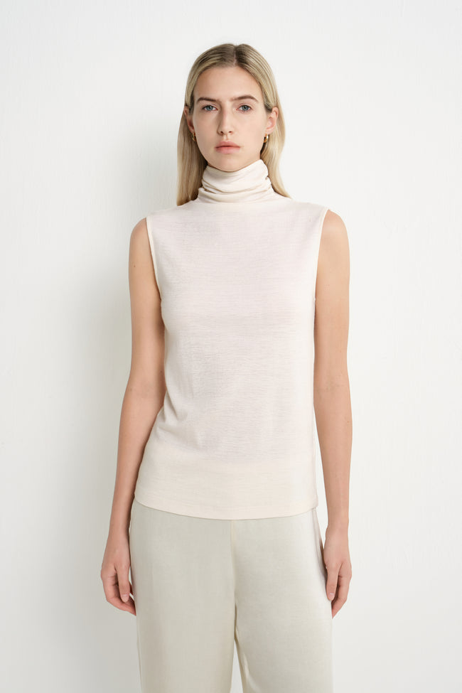 A blonde model wearing a sleeveless, ribbed-knit turtleneck top in a warm off-white/cream color, paired with light beige trousers, against a plain white studio background.