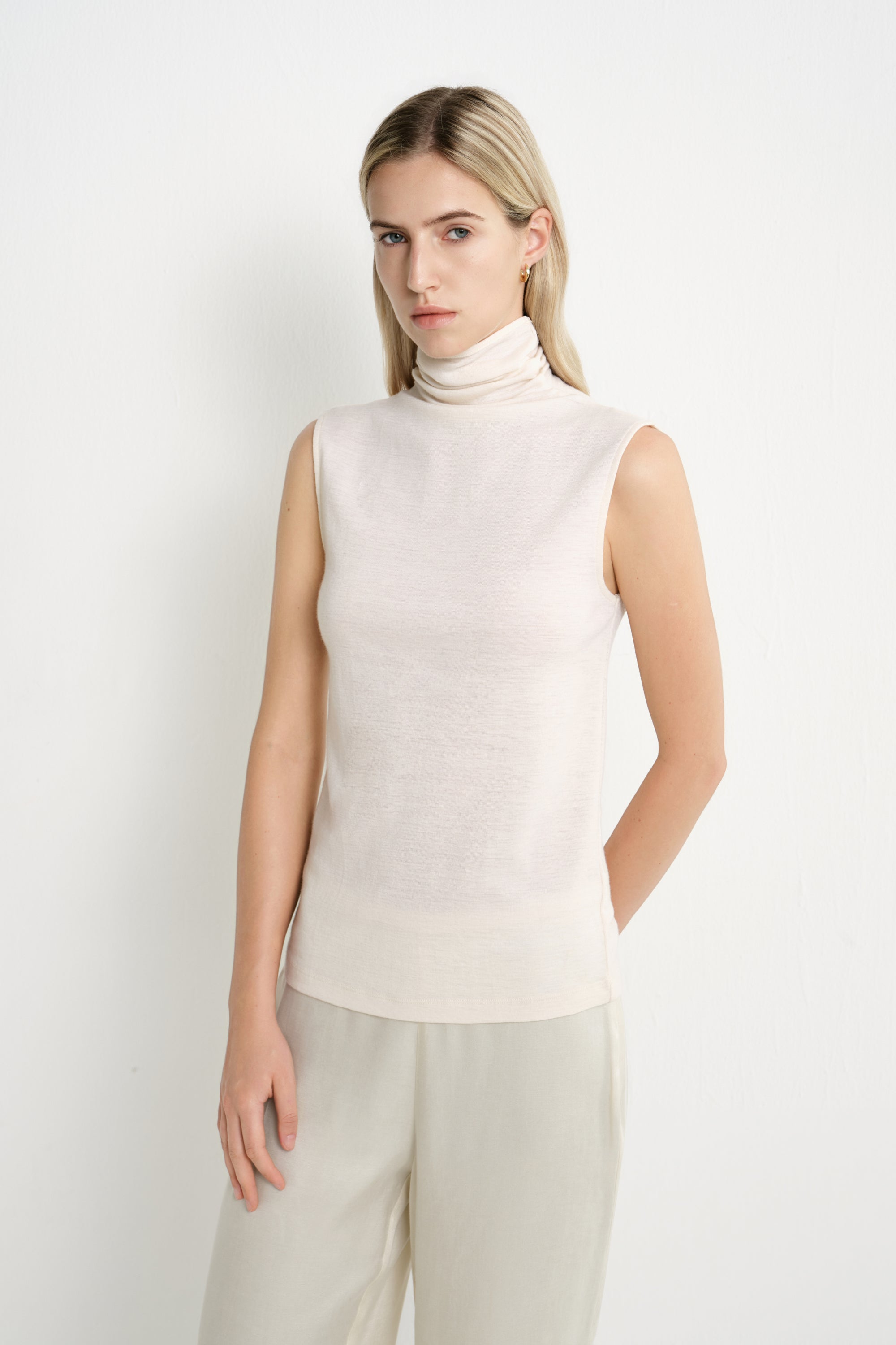 A model wearing a Rue Sophie creme sleeveless mock-neck top with a draped high neckline and subtle textured finish, three-quarter front view.
