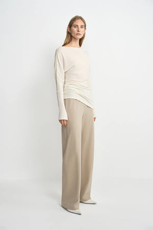 A model wearing oatmeal wide-leg wool-blend trousers with soft fluid drape, side angle view, pairing an ivory boat-neck top and white pointed-toe heels.