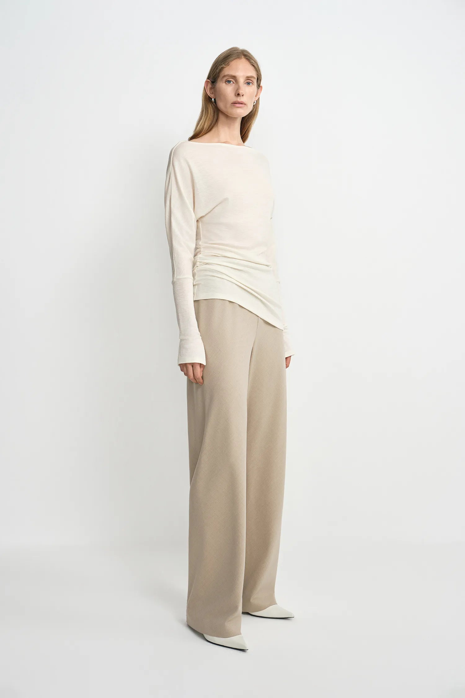 A model wearing oatmeal wide-leg wool-blend trousers with soft fluid drape, side angle view, pairing an ivory boat-neck top and white pointed-toe heels.