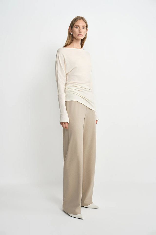 A model wearing oatmeal wide-leg wool-blend trousers with soft fluid drape, side angle view, pairing an ivory boat-neck top and white pointed-toe heels.