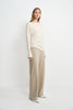 A model wearing oatmeal wide-leg wool-blend trousers with soft fluid drape, side angle view, pairing an ivory boat-neck top and white pointed-toe heels.