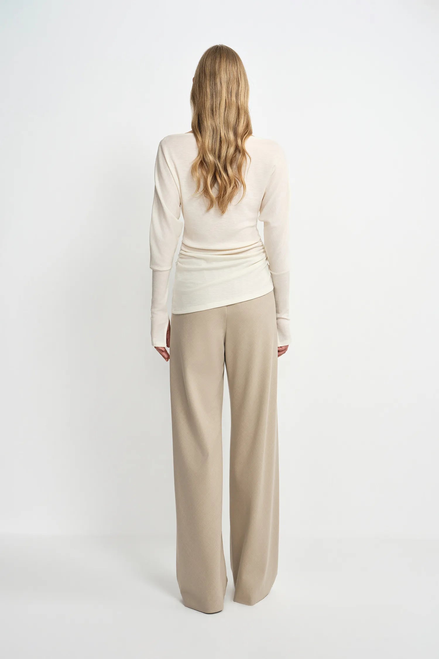 A model wearing oatmeal wide-leg wool-blend trousers, back view showing the relaxed silhouette and fluid drape, pairing an ivory draped top.