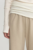 Close-up of a person wearing light beige wide-leg trousers and an off-white long-sleeve top, with the hand resting near the trouser leg against a plain light background.