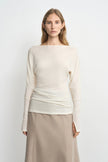 A woman modeling a cream-colored knit long-sleeve top with a wide boat neckline, paired with a beige skirt, against a plain light gray background.