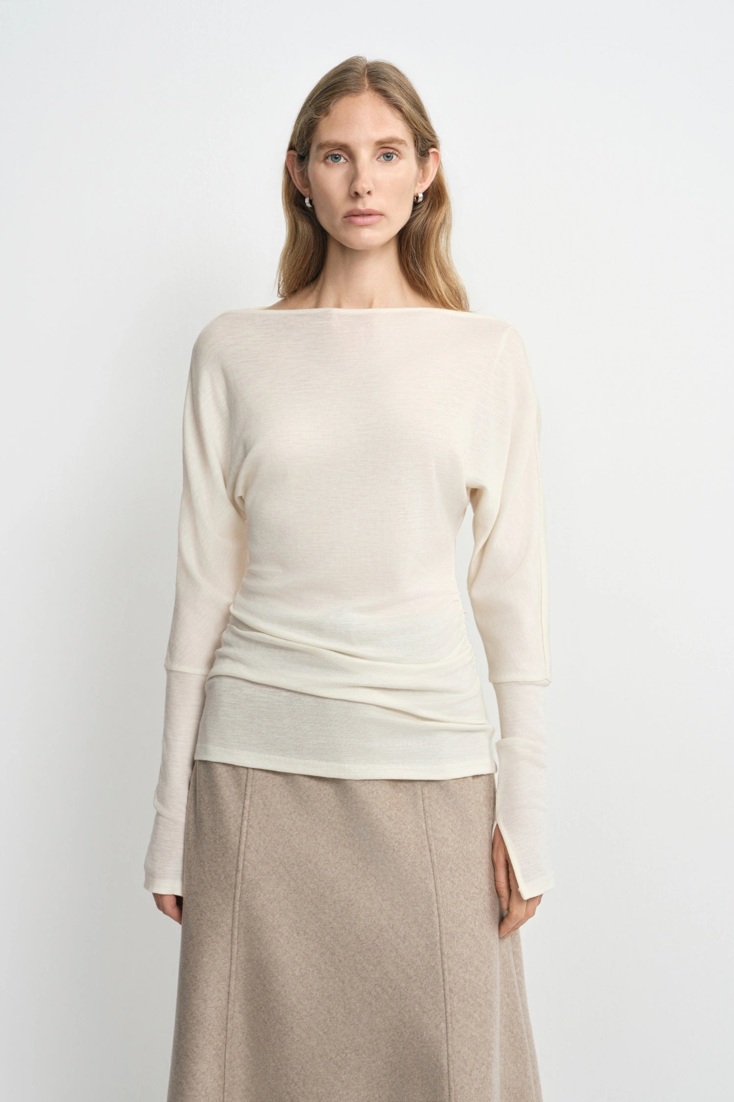 A woman modeling a cream-colored knit long-sleeve top with a wide boat neckline, paired with a beige skirt, against a plain light gray background.