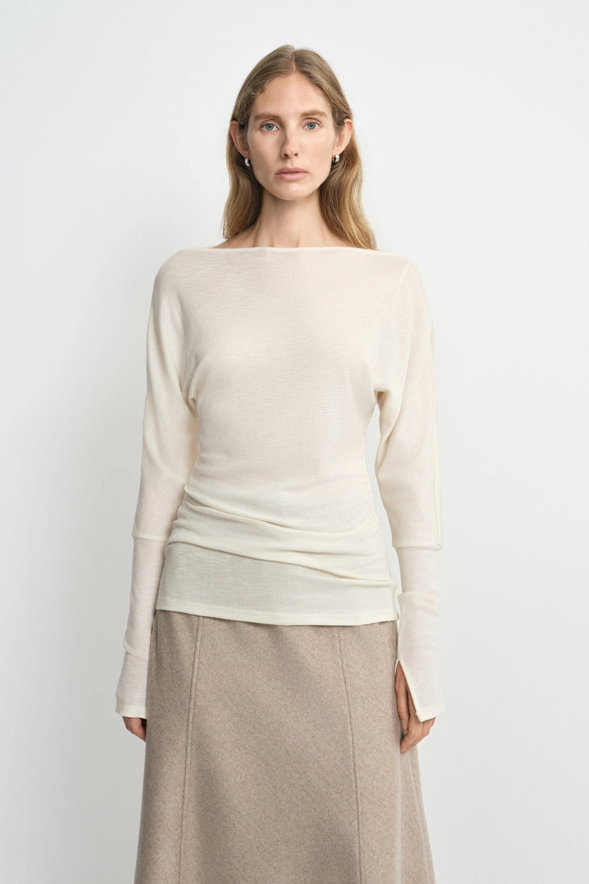 A woman modeling a cream-colored knit long-sleeve top with a wide boat neckline, paired with a beige skirt, against a plain light gray background.