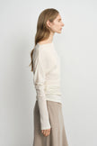 A woman shown in side profile wearing a cream long-sleeve knit top with ruching and a beige skirt, photographed against a plain light gray studio background.
