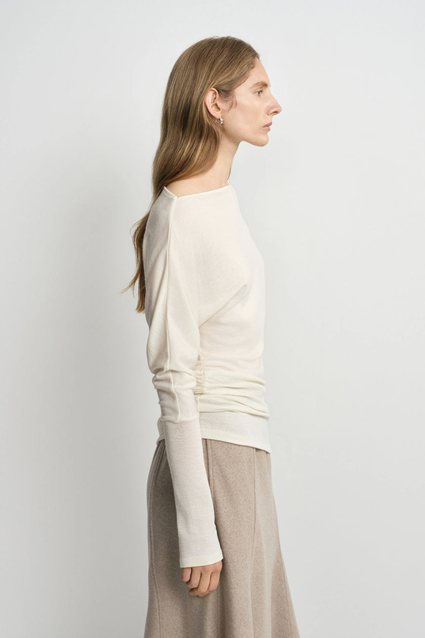 A woman shown in side profile wearing a cream long-sleeve knit top with ruching and a beige skirt, photographed against a plain light gray studio background.