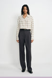 A model stands against a light gray studio background wearing a beige and white plaid button-down shirt tucked into dark charcoal gray wide-leg trousers, paired with a black belt and black shoes.