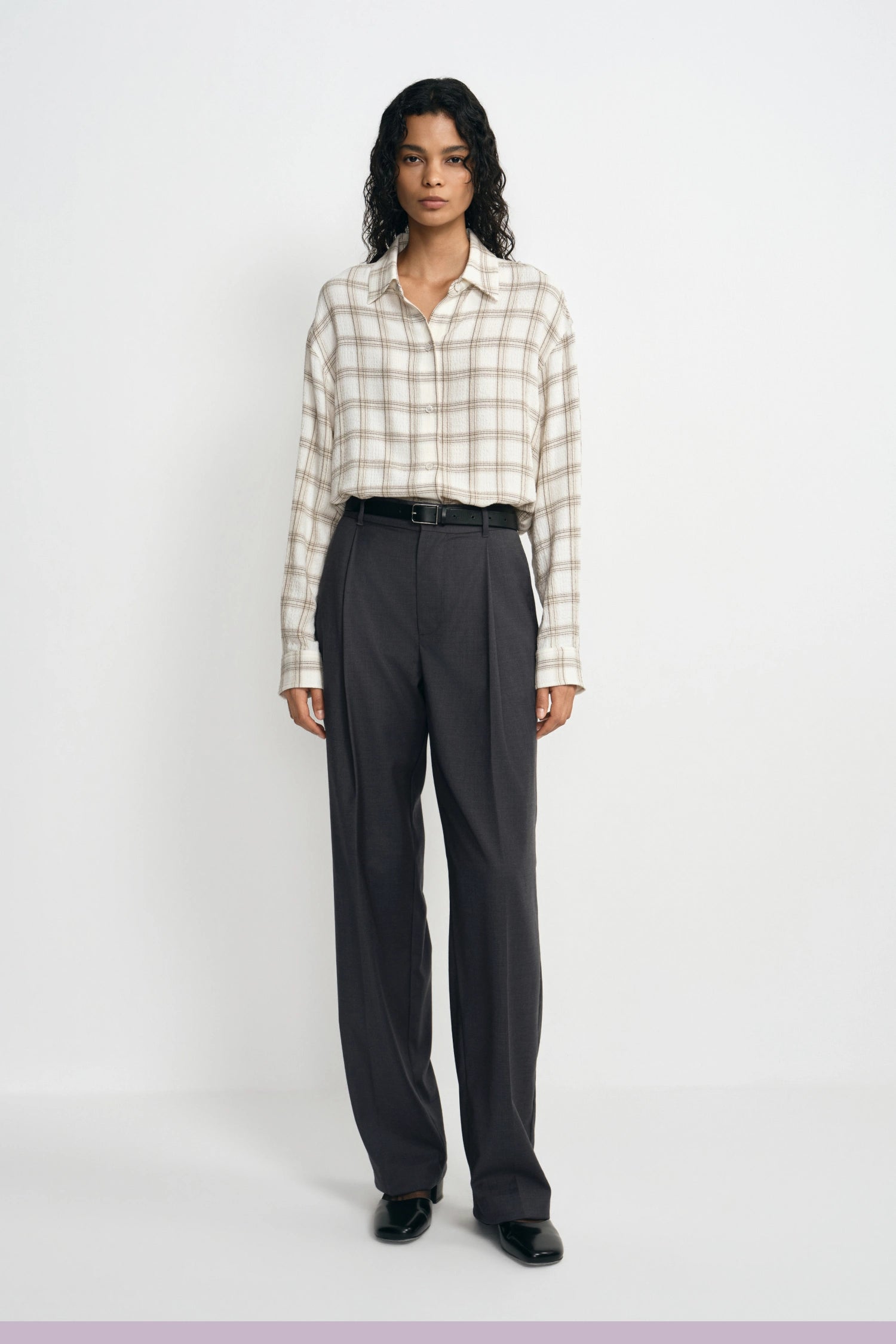 A model stands against a light gray studio background wearing a beige and white plaid button-down shirt tucked into dark charcoal gray wide-leg trousers, paired with a black belt and black shoes.