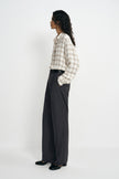 A model is shown from a side profile wearing a beige and white plaid button-down shirt tucked into dark charcoal gray wide-leg trousers, paired with black shoes, against a plain white studio background.