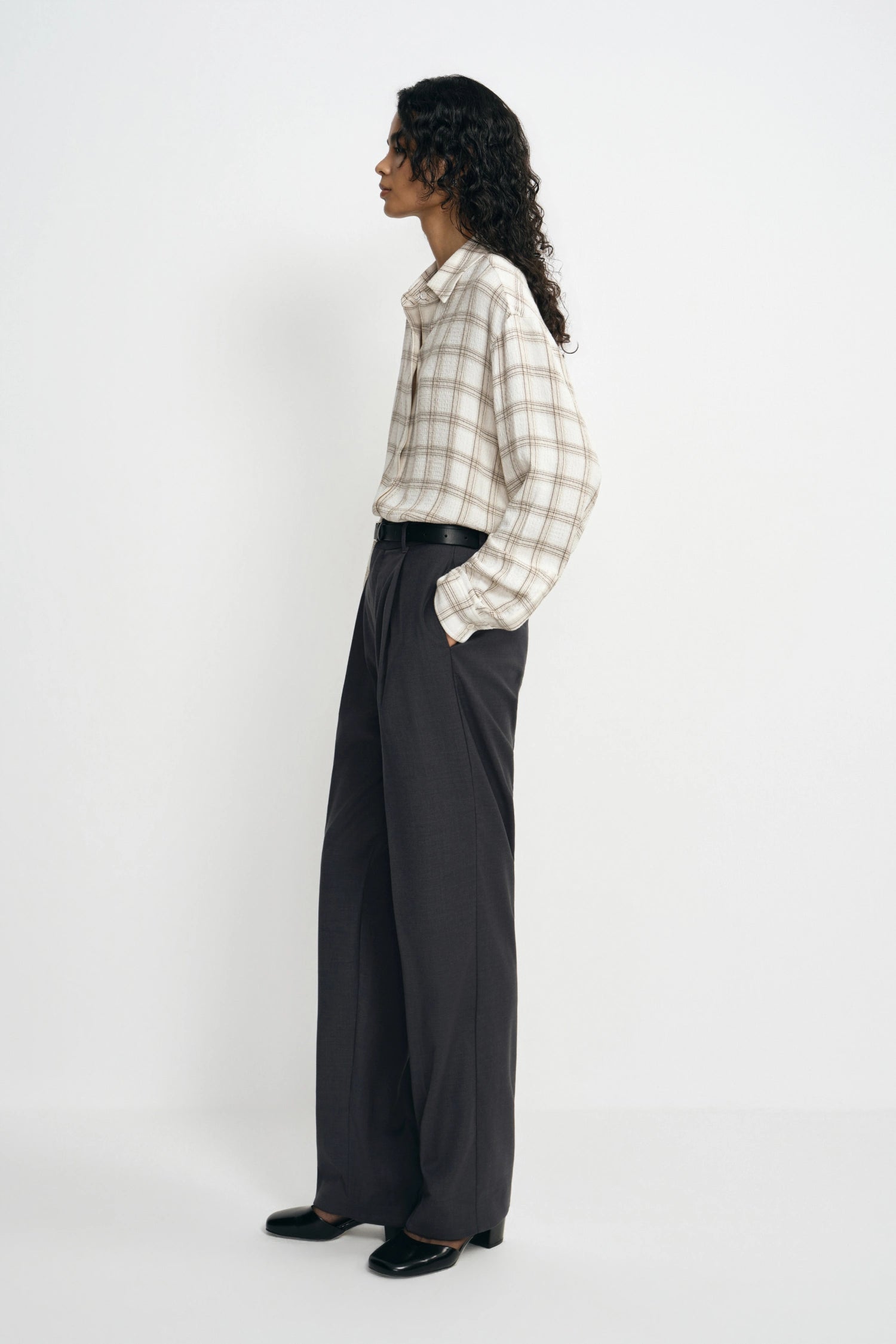 A model is shown from a side profile wearing a beige and white plaid button-down shirt tucked into dark charcoal gray wide-leg trousers, paired with black shoes, against a plain white studio background.